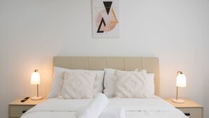A neatly made bed with white linens and pillows, two bedside lamps, and a wall-mounted abstract art piece. - Modern 1BR Apartment | Convenient Stay (MacGregor)
