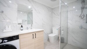 A modern bathroom with a marble floor, a glass shower enclosure, a white toilet, and a washing machine. - Modern 1BR Apartment | Convenient Stay (MacGregor)