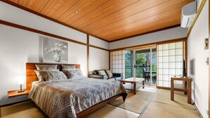 Een traditionele Japanse slaapkamer met tatami vloeren, een bed met beddengoed met een patroon, een televisietoestel en een glazen schuifdeur naar een balkon.