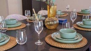 Een eettafel gedekt met meerdere borden, kommen en wijnglazen, met een vaas en stokjes op tafel.
