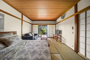 Une pièce de style japonais traditionnel, avec un sol recouvert de tatamis, un plafond bas en bois, un lit avec une literie à motifs, un canapé, une petite table et des portes coulissantes donnant sur un espace extérieur.