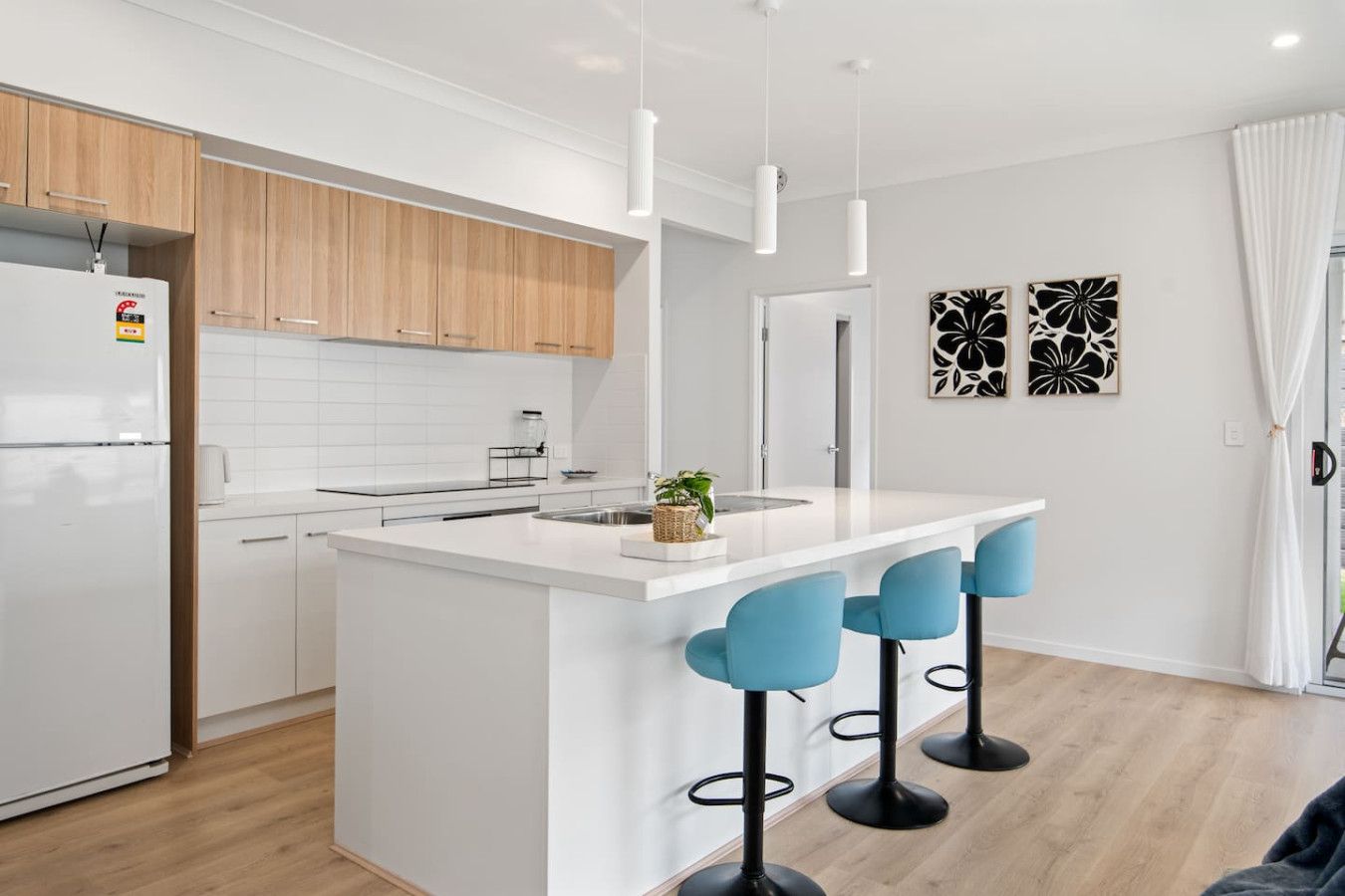 Een moderne keuken met witte kasten, een centraal eiland met barstoelen, hanglampen en uitzicht op de woonkamer.