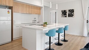 A modern kitchen with white cabinetry, a central island with bar stools, pendant lighting, and a view of the living area.