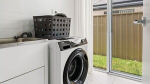 A laundry room with a front-loading washer and dryer, a gray laundry basket, and a window with a view of a backyard.
