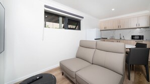 En moderne stue med en beige sofa, et lille spisebord med stole, et køkkenområde med træskabe og et vindue med udsigt.