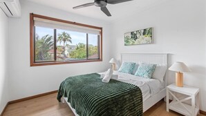 A bedroom with a bed, a ceiling fan, a window with a view of palm trees, and a painting on the wall. - Elegant 4BR Residence |Friendly Haven in Calamvale (Calamvale)