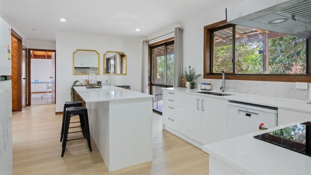 A modern kitchen with a central island, white cabinetry, and a large window providing a view of greenery. - Elegant 4BR Residence |Friendly Haven in Calamvale (Calamvale)