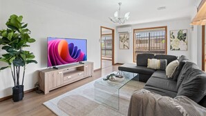 A modern living room with a flat-screen TV, a grey sofa, a glass coffee table, and a large plant. - Elegant 4BR Residence |Friendly Haven in Calamvale (Calamvale)