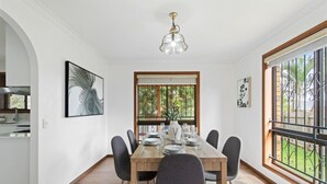 A dining area with a wooden table set for six, surrounded by chairs. There is a window with a view of greenery outside, a kitchen visible to the left, and a ceiling light fixture above the table. - Elegant 4BR Residence |Friendly Haven in Calamvale (Calamvale)