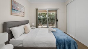 A bedroom with a bed, bedside table, lamp, and a view of an outdoor area through a sliding door. - Charming 2BR Home in Serene Mount Warren Park (Mount Warren Park)