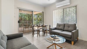 A living room with a grey sofa, a glass coffee table, a dining table with chairs, and a sliding glass door leading to an outdoor area. - Charming 2BR Home in Serene Mount Warren Park (Mount Warren Park)