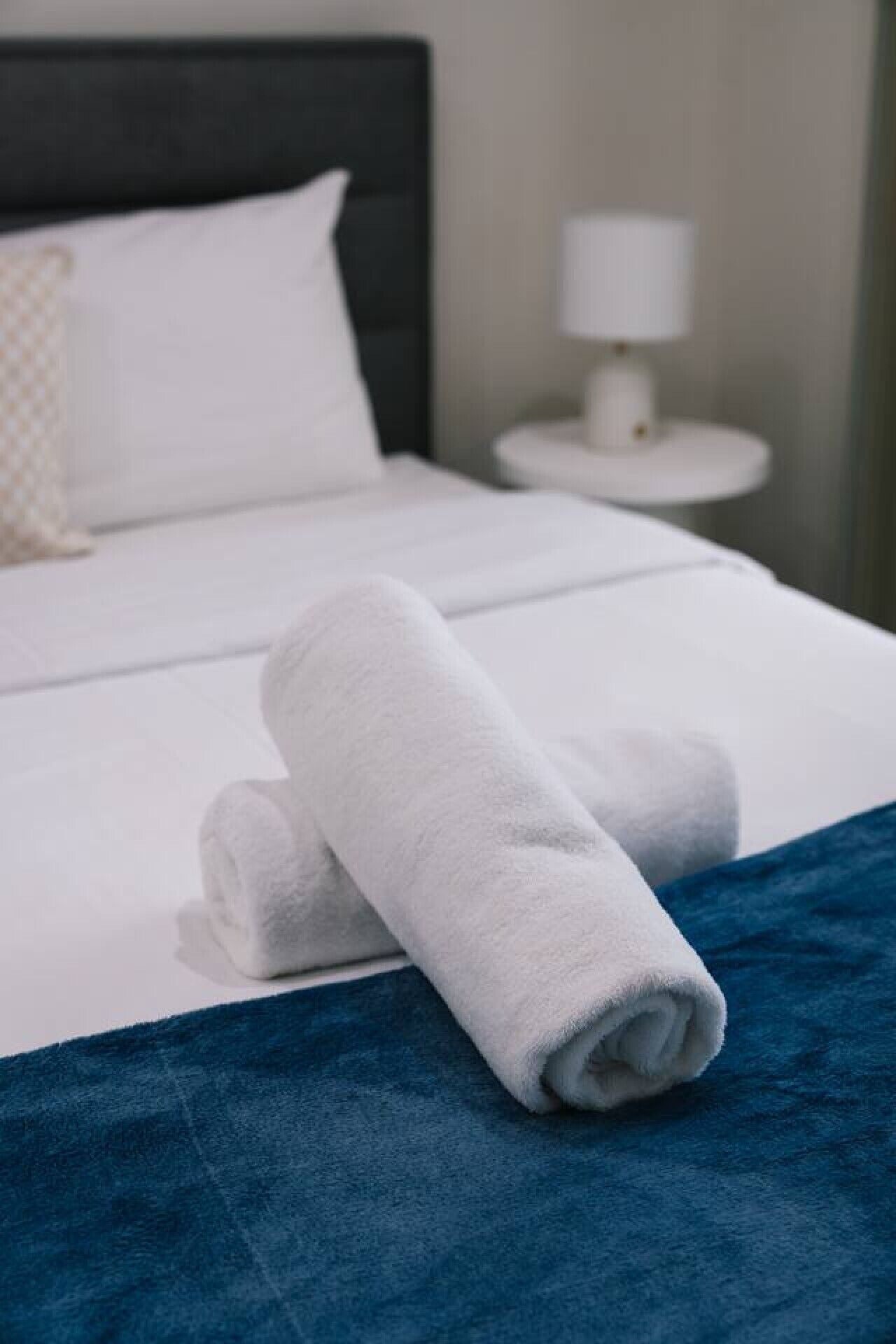 Een net opgemaakt bed met witte lakens, een opgerolde witte handdoek op het bed en een blauw accent op de spreideken.