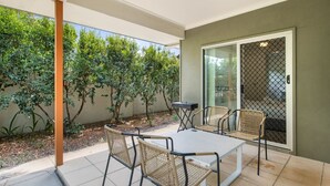 A patio with a table and chairs, surrounded by greenery and a sliding glass door.