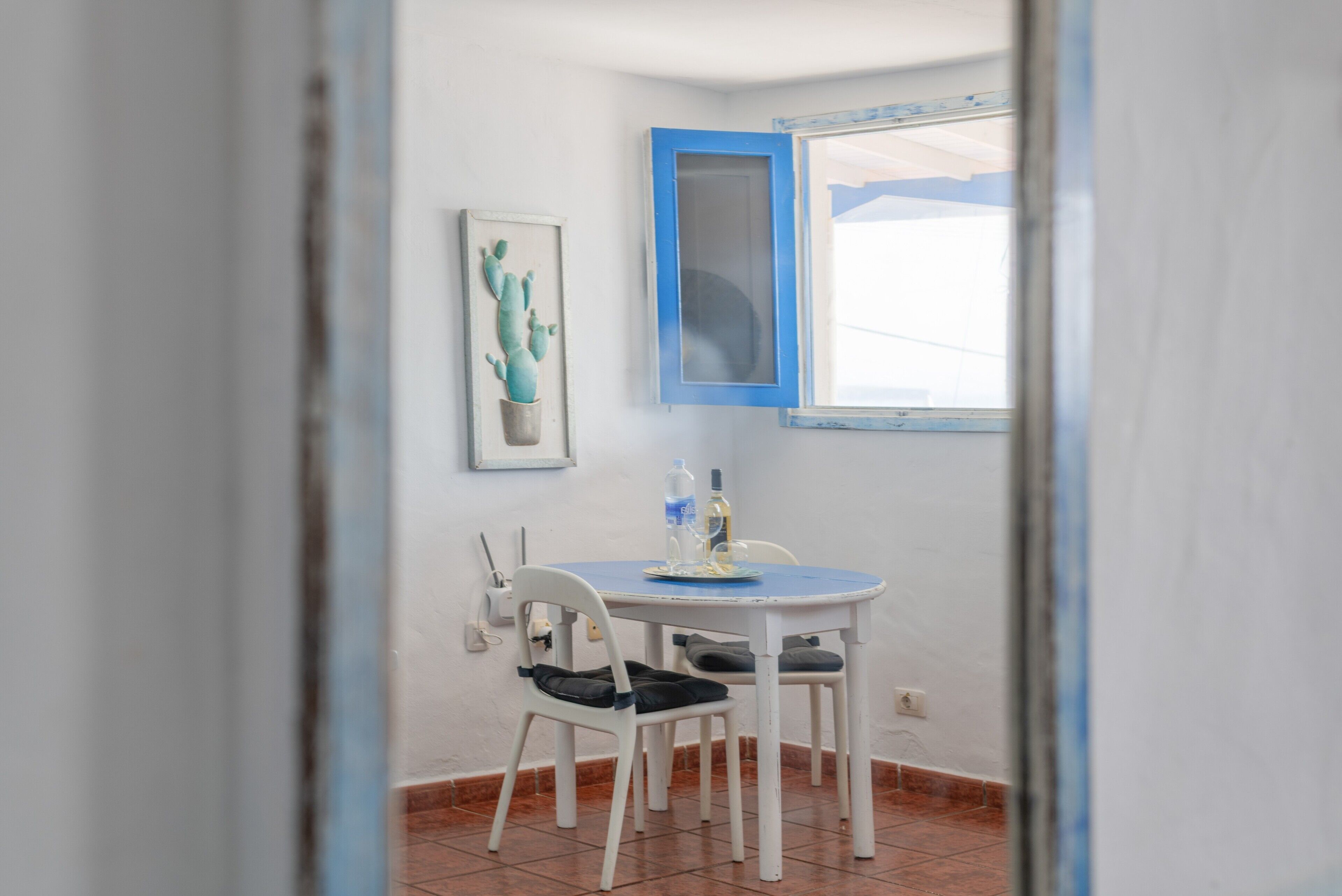 Un coin repas avec une table ronde, des chaises, une décoration en forme de cactus et une fenêtre à cadre bleu.