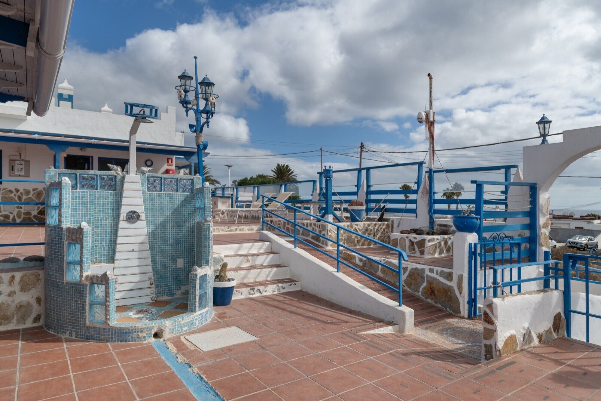Un paseo marítimo con barandillas azules, una fuente de azulejos y vista al mar.