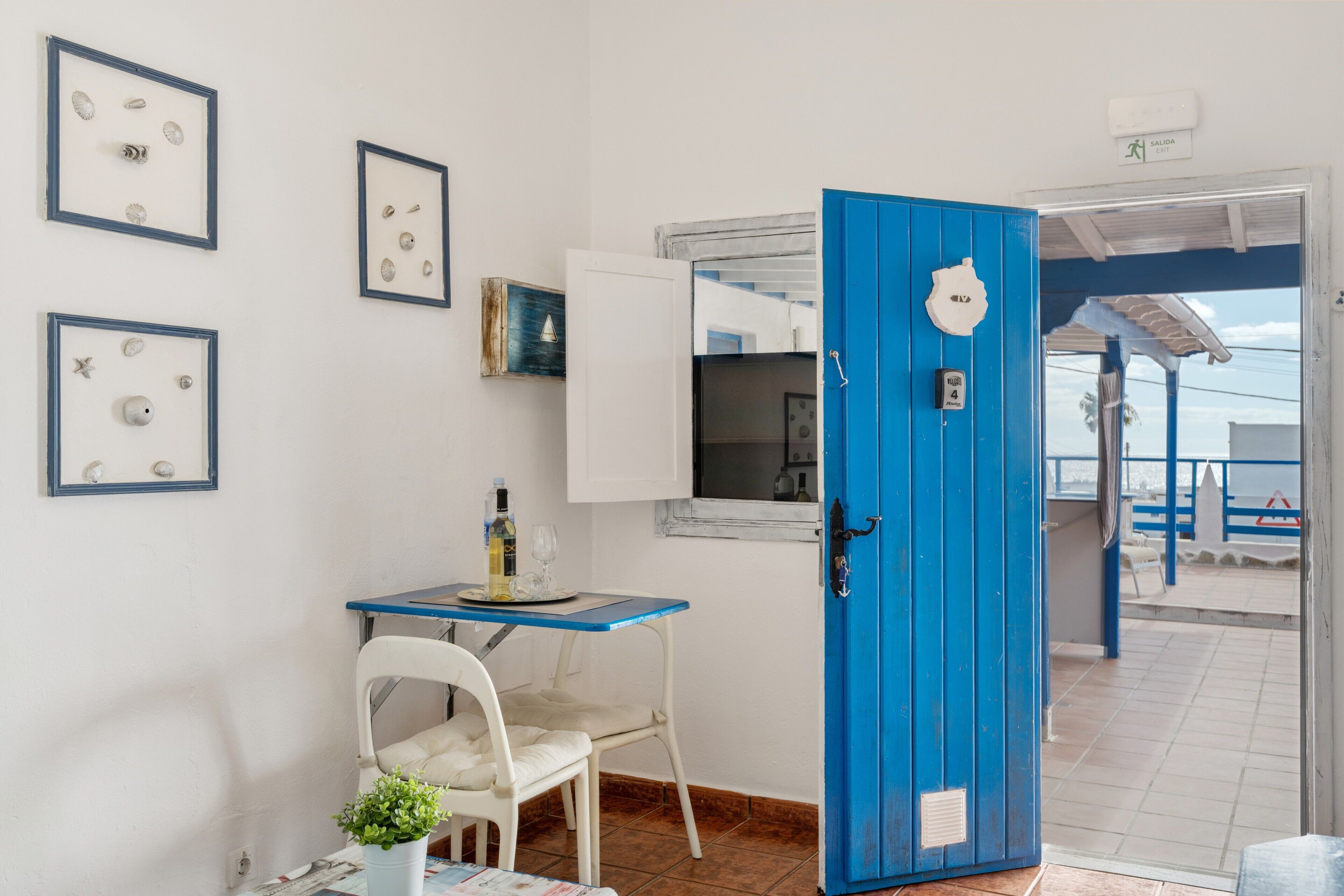 A room with a blue door, white walls, framed pictures, a small table with a chair, and a view of the outside.