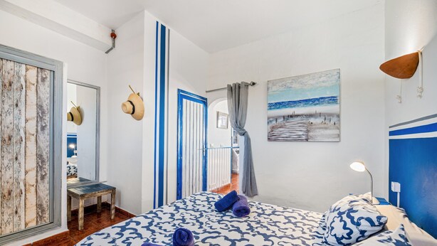 Ein Schlafzimmer mit einem gemusterten Bettüberwurf, einem Strandbild und Meerblick durch ein Fenster.