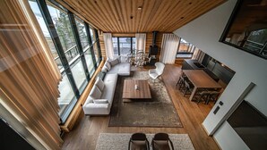 Ein modernes Interieur mit Holzboden, großen Fenstern, einem Ecksofa, einem Esstisch und einer Küchenzeile.
