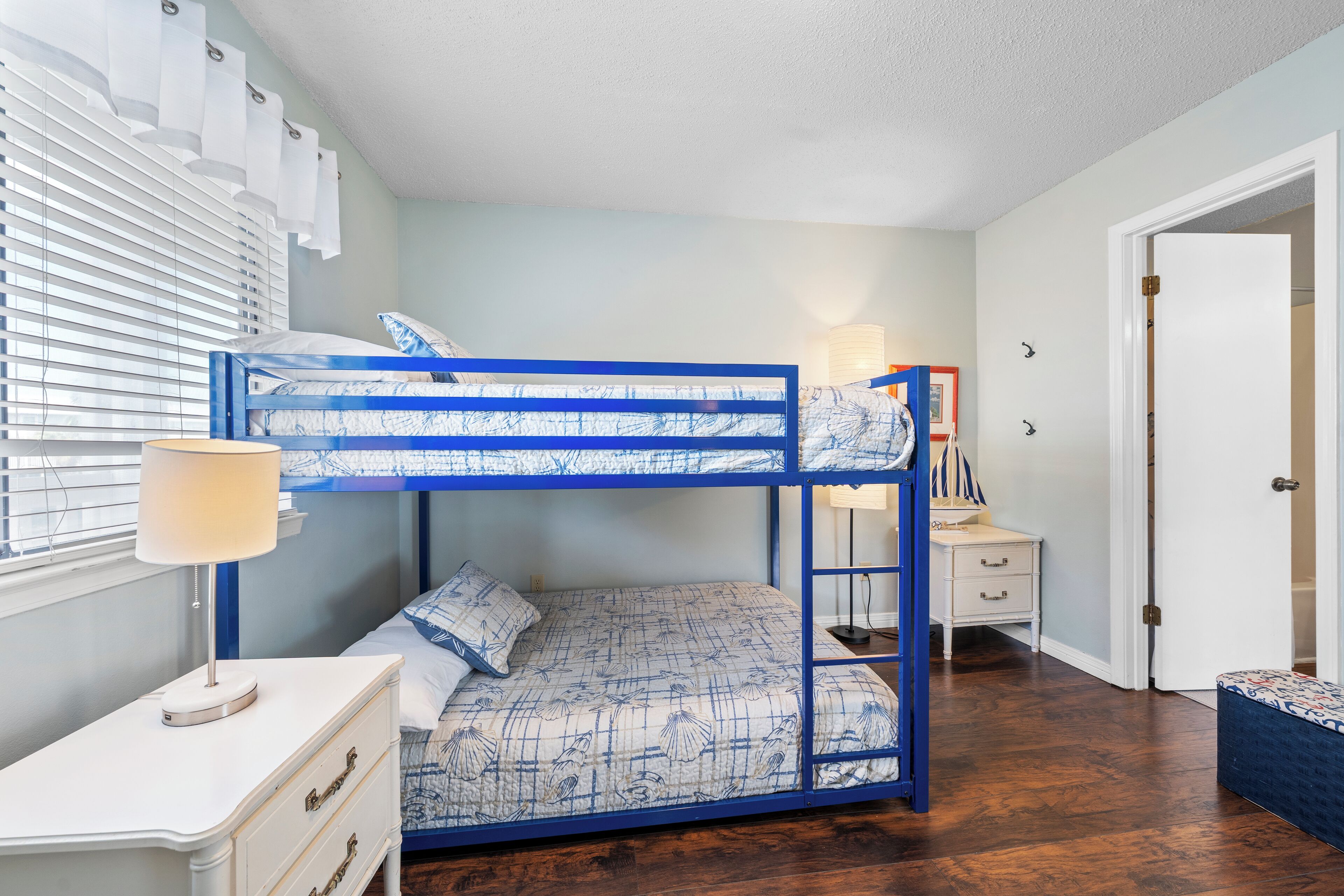 A bunk bed with a blue frame and patterned bedding, a bedside table with a lamp, a white dresser, and a door leading to another room.