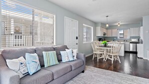 A modern living room with a grey sofa, dining table, and chairs. There is a kitchen area with white cabinets and a microwave. - Wolf Bay Villas 208 (Orange Beach)