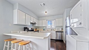 A kitchen with white cabinets, a central island with stools, stainless steel appliances, and a window with blinds. - Wolf Bay Villas 208 (Orange Beach)