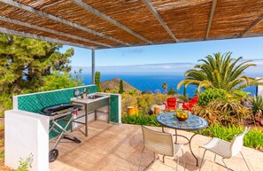 Um pátio externo com churrasqueira, uma mesa com frutas e vista para o oceano.