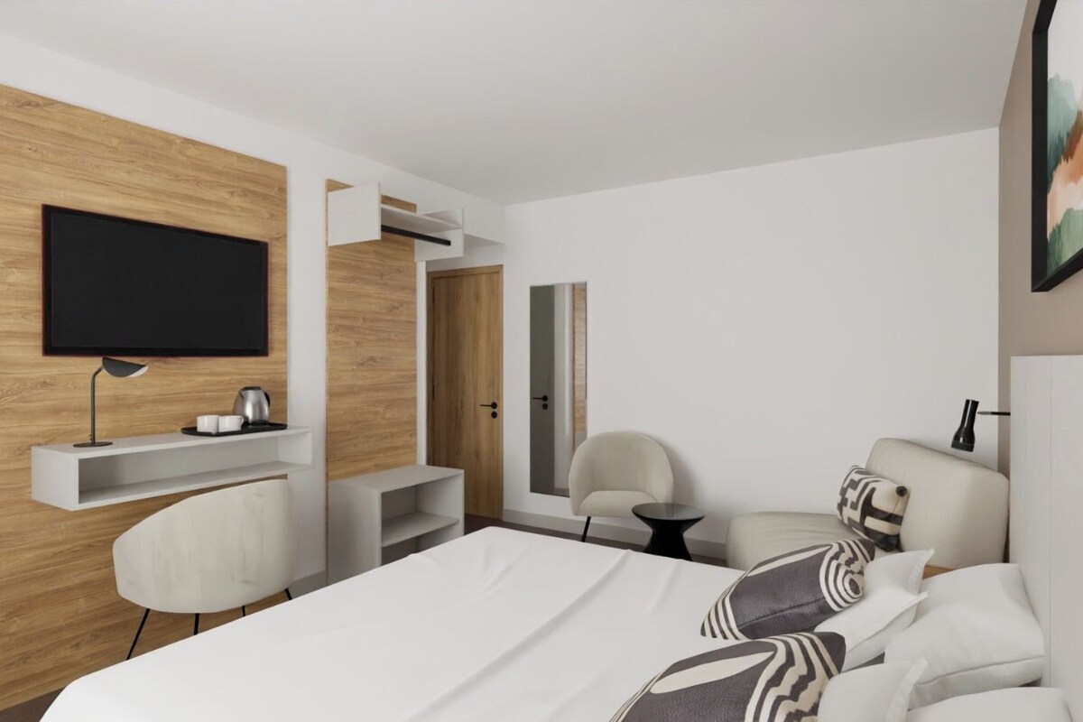 A modern hotel room with a large bed, a flat-screen TV mounted on the wall, a desk with a chair, and a small table.