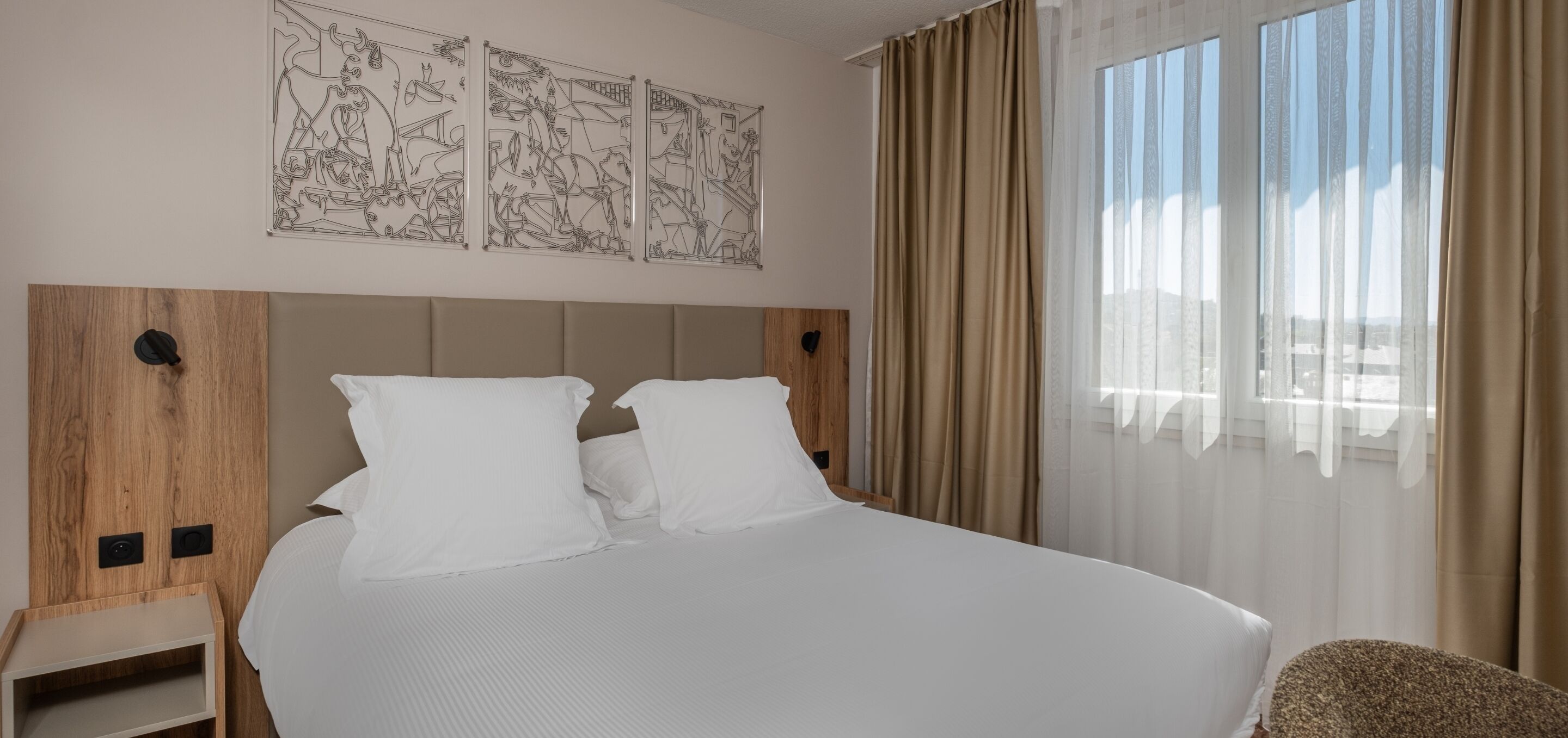 A hotel room with a large bed, a wooden headboard, a nightstand, and a view of the city through sheer curtains.