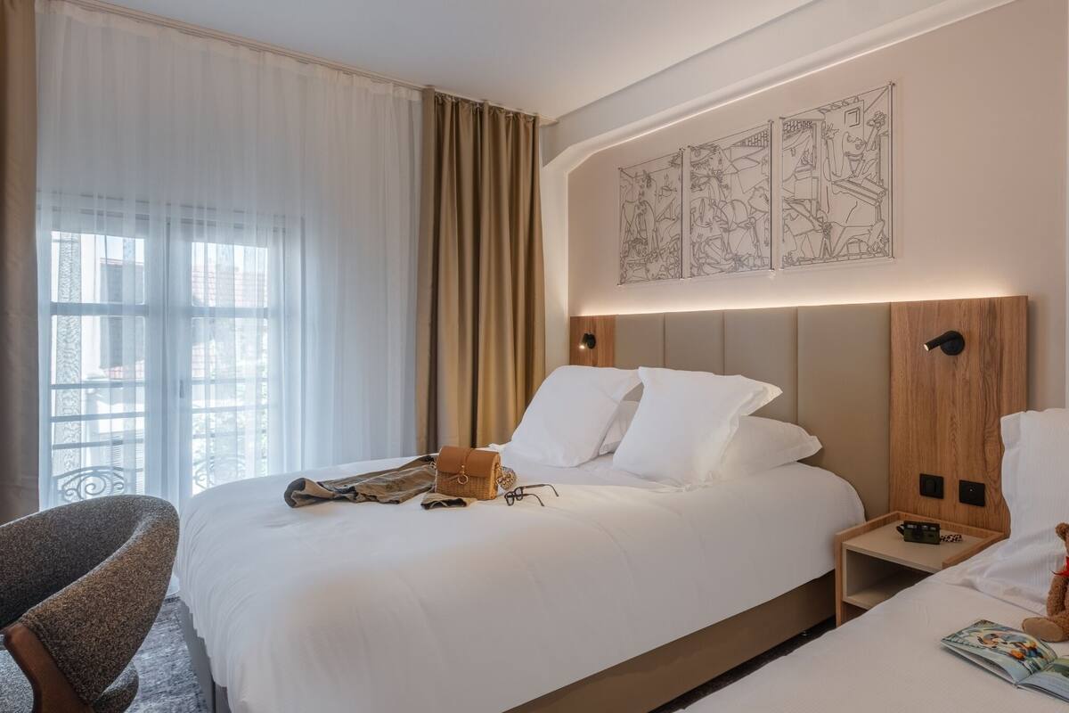 A hotel room with a bed, bedside table, chair, and a teddy bear.