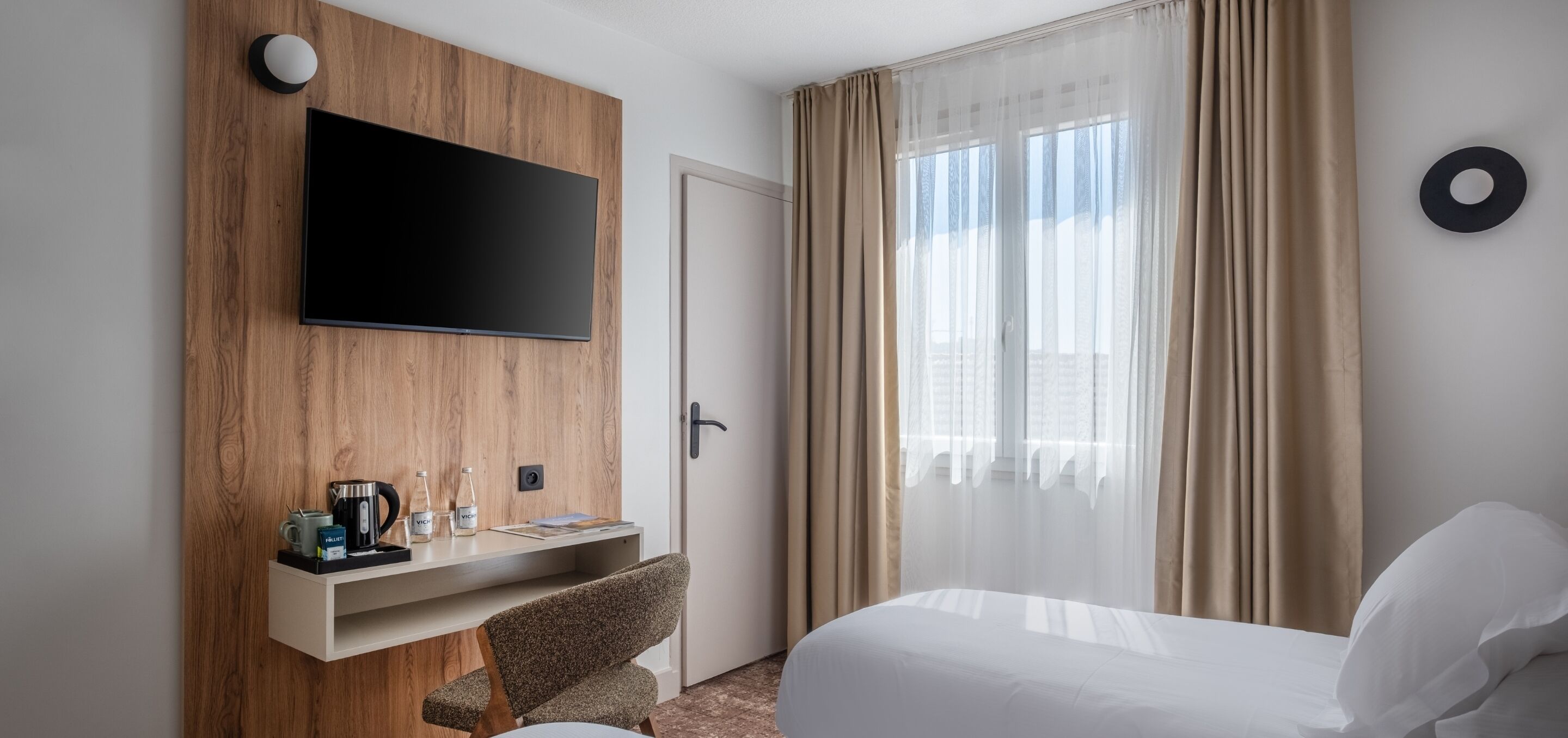 Habitación de hotel con televisor de pantalla plana montado en un panel de madera, un escritorio con cafetera y botellas, una silla, una cama con ropa blanca y un ventanal con cortinas translúcidas.
