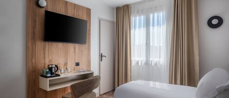 Habitación de hotel con televisor de pantalla plana montado en un panel de madera, un escritorio con cafetera y botellas, una silla, una cama con ropa blanca y un ventanal con cortinas translúcidas.