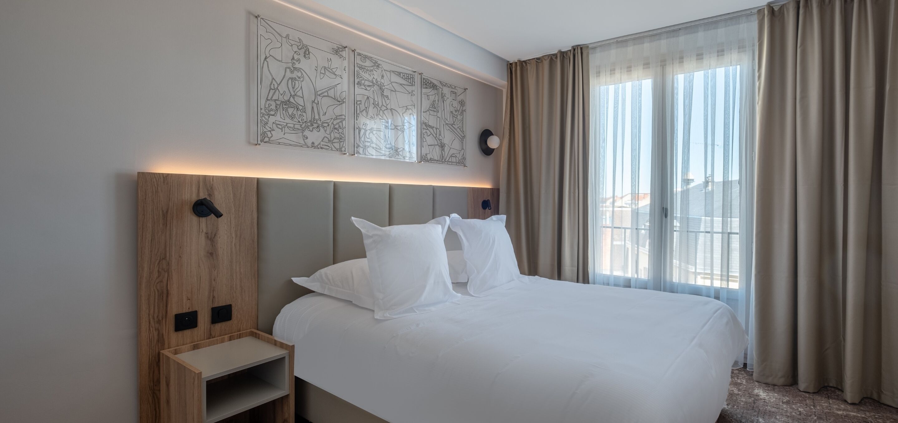 Habitación de hotel con una cama grande, una mesita de noche, cabecera con obra de arte y vistas al exterior a través de cortinas translúcidas.