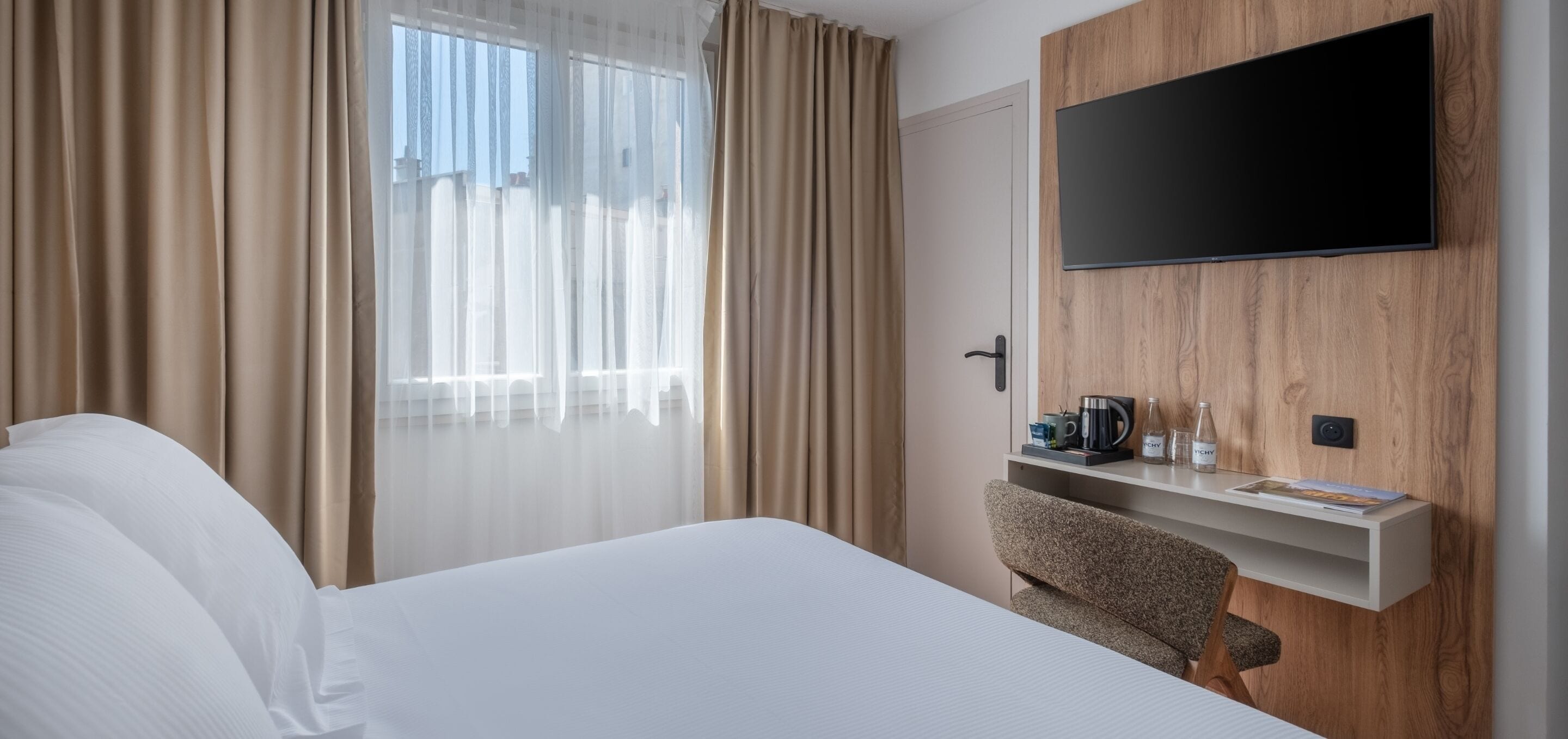 Et hotelværelse med en seng, et fjernsyn ophængt på en trævæg, et lille bord med kaffemaskine og en stol.