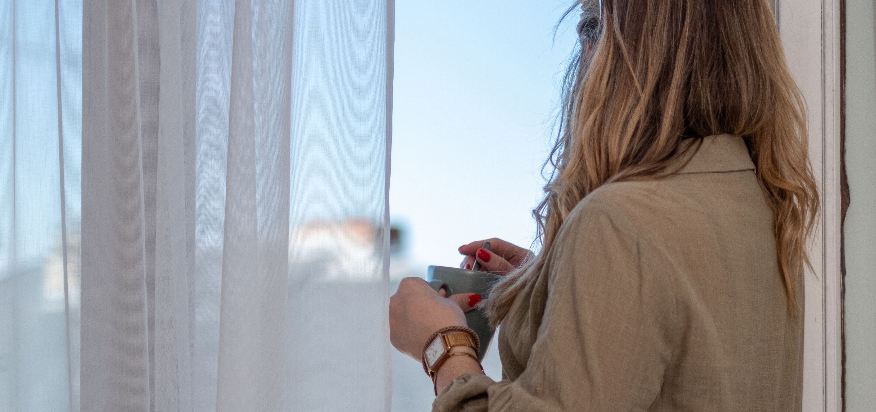 A person holding a cup near a window with sheer curtains.