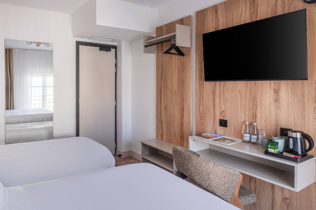 A hotel room with a bed, a TV mounted on a wooden wall, a bedside table with a kettle, and a mirror.