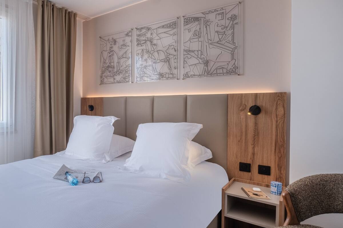 A hotel room with a large bed, a bedside table, a chair, and a wall with artwork.
