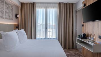Ett hotellrum med en säng, gardiner, en väggmonterad TV och en liten hylla med diverse föremål.
