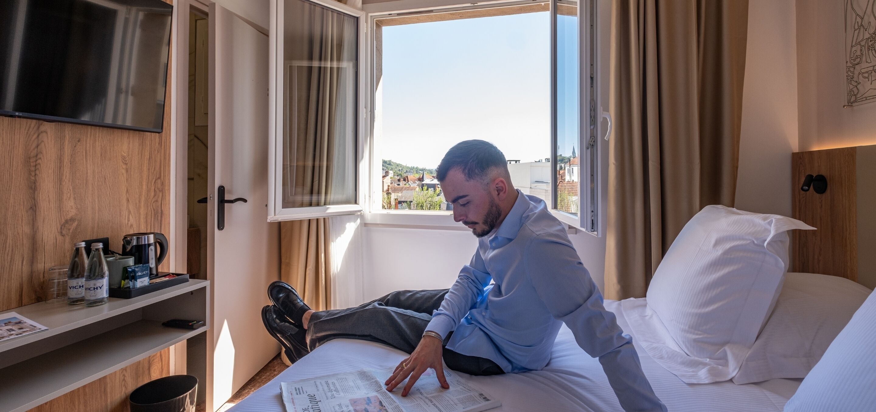 Un homme est assis sur un lit, lisant un journal, avec une fenêtre qui donne sur l’extérieur.