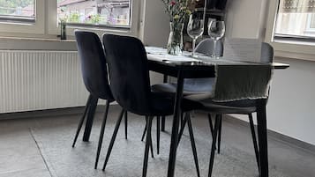 Un coin repas comprenant une table ronde, quatre chaises, un vase avec des fleurs et une fenêtre donnant sur des maisons.