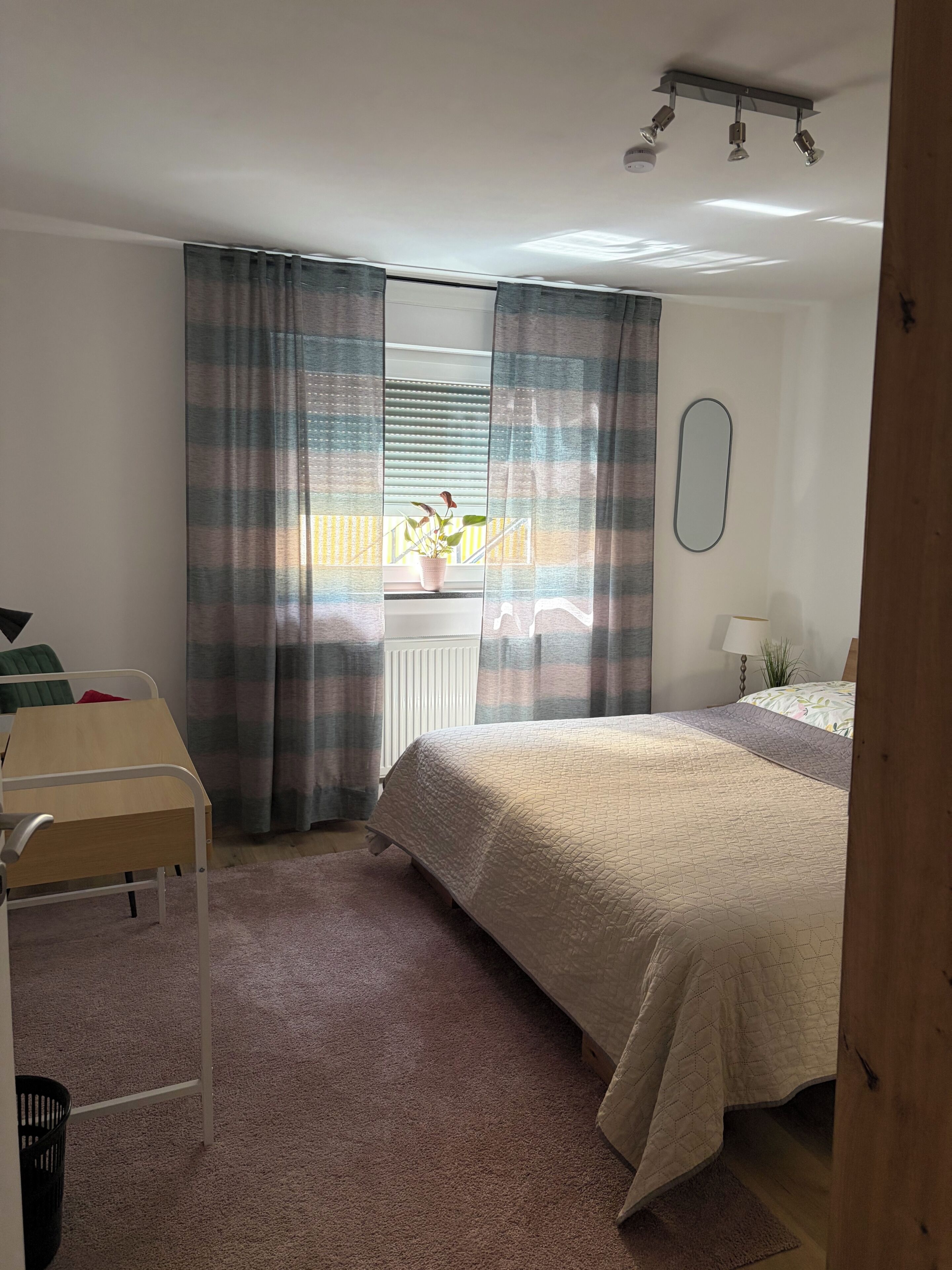 A bedroom with a bed, a desk, a chair, a radiator, and a window with curtains.