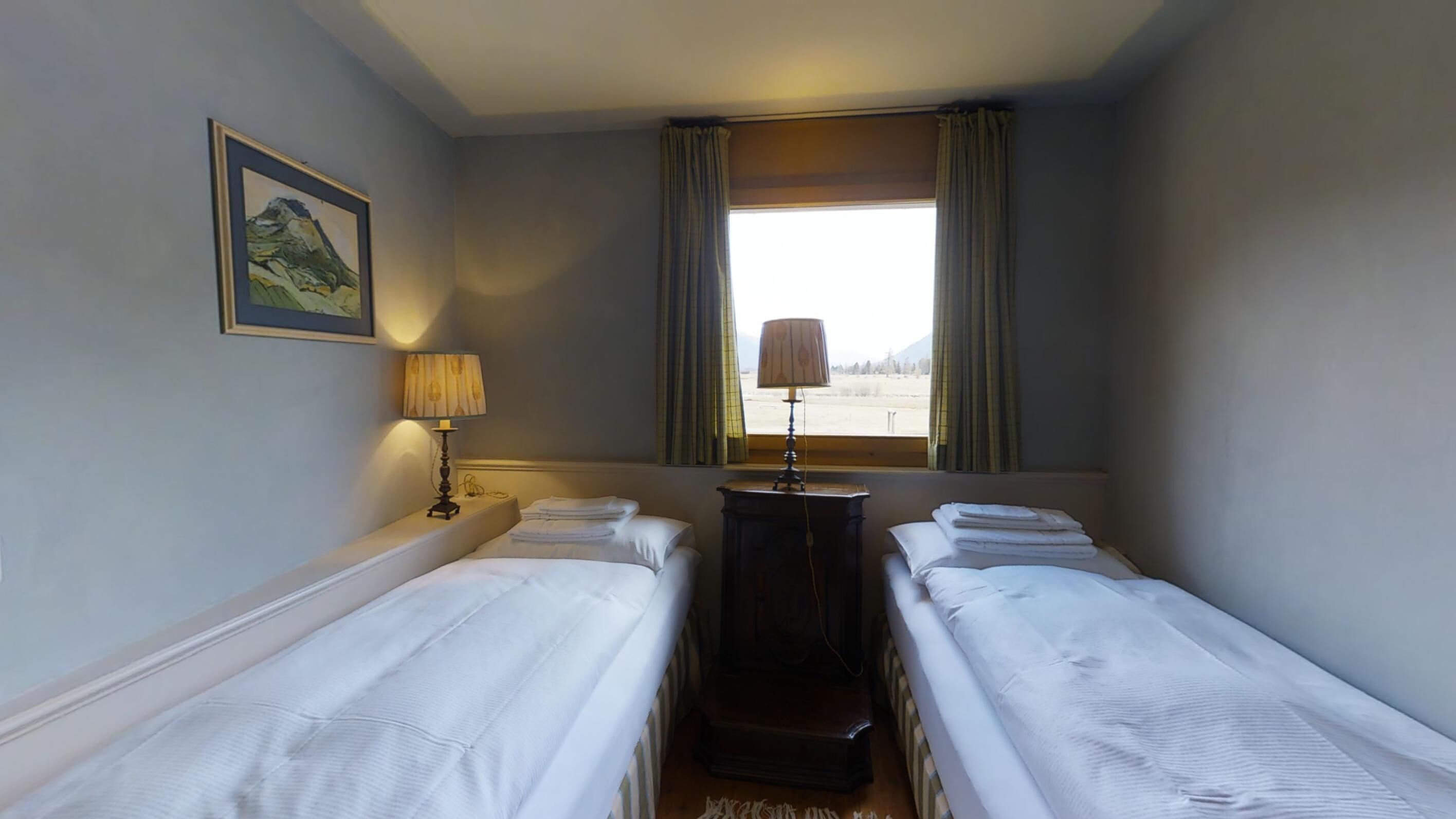 Quarto de hotel com duas camas de solteiro, mesa de cabeceira com abajur, quadro na parede e janela com cortinas.