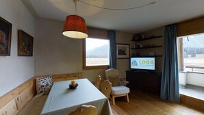 A cozy room with a wooden table, a bench, a television displaying 'LOGA', and a window with a view of a snowy landscape. - 3-room apartment approx. (Celerina/Schlarigna)