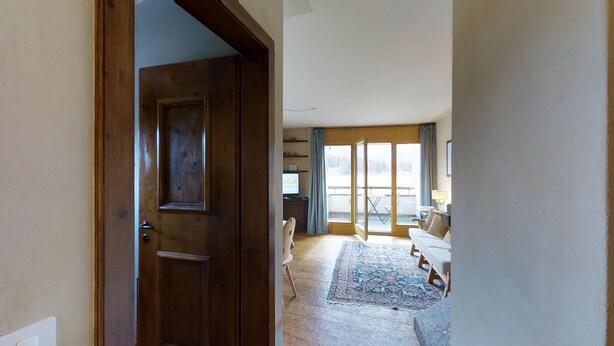 A room with a wooden door, a sofa, a patterned rug, and a view through a window. - 3-room apartment approx. (Celerina/Schlarigna)