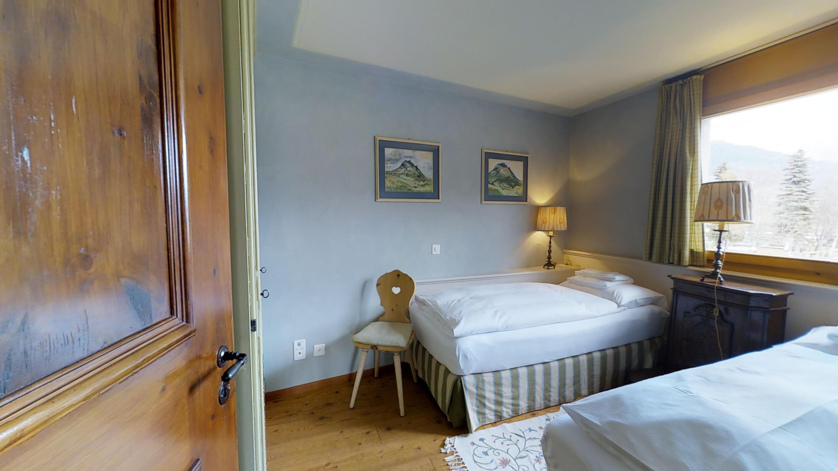 Un dormitorio con dos camas, una puerta de madera, una silla, una lámpara y dos cuadros en la pared.