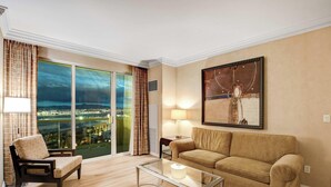 Camera d'albergo con un'ampia finestra che si affaccia sulla città, un divano, una poltrona, una lampada e un quadro appeso al muro.