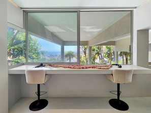 A modern room with a large window, a white desk, two chairs, and a view of a cityscape and greenery. - The White Camel Villa (Cape Town)