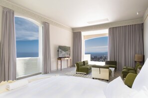 Habitación de hotel con una cama grande, dos sillones verdes, un sofá blanco, un cuadro en la pared y vistas a la ciudad a través de amplias ventanas.