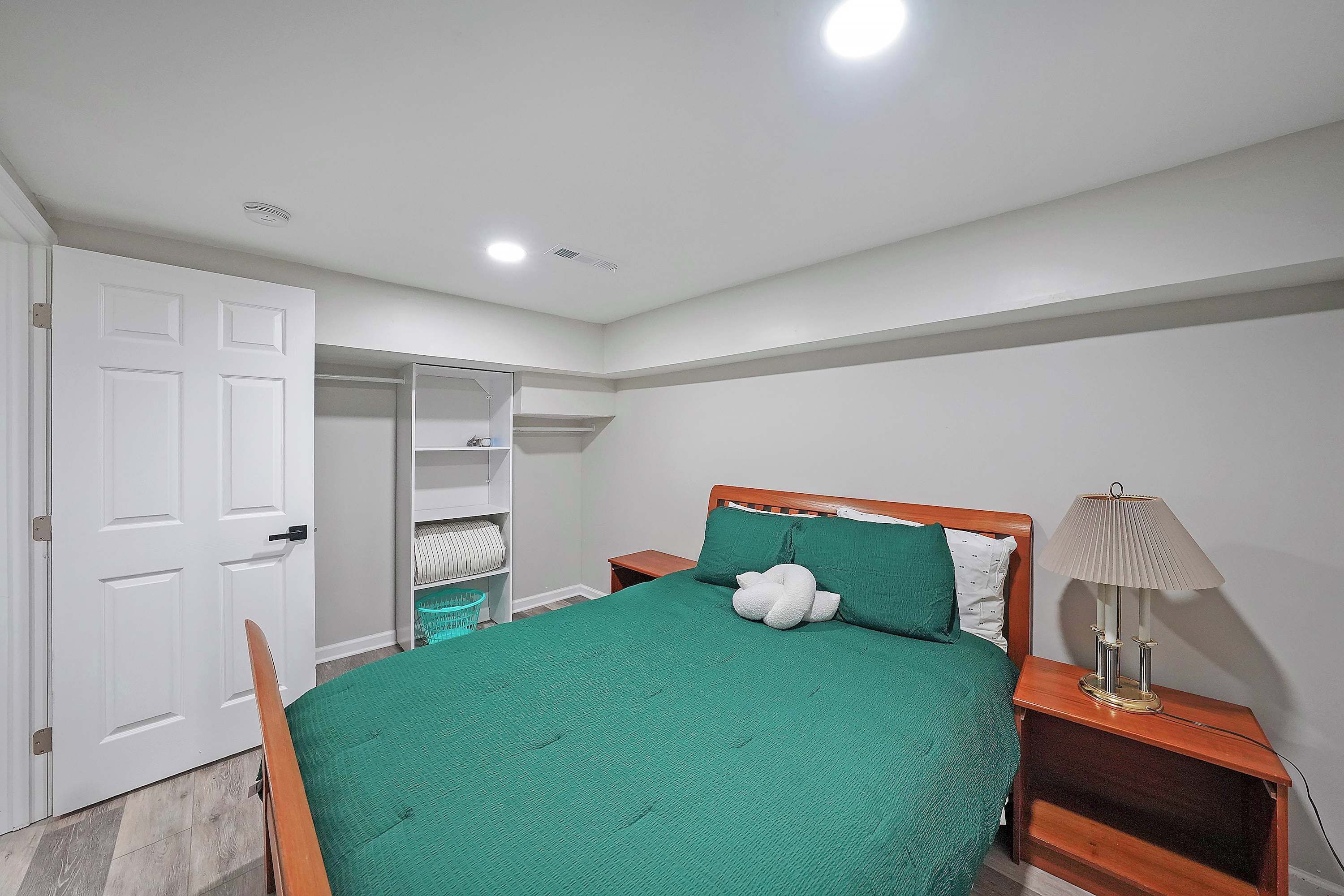 A bedroom with a green bedspread, a wooden nightstand with a lamp, a white door, and a closet with shelves.