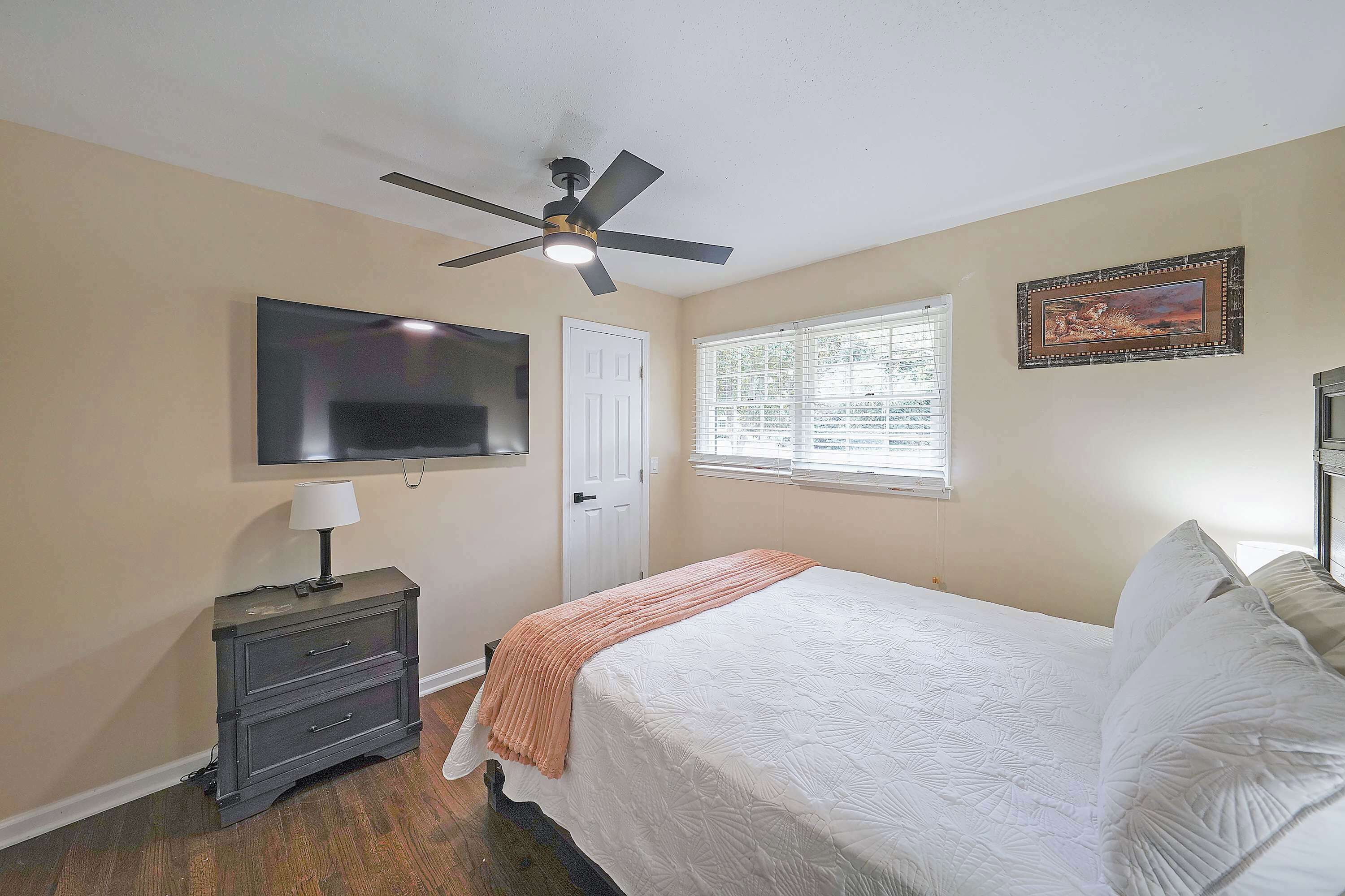 A bedroom with a bed, a nightstand, a TV, a ceiling fan, a window, and a door.