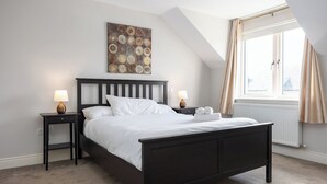 Ein Schlafzimmer mit einem dunklen Holzbett, weißer Bettwäsche, zwei Nachttischen mit Lampen, einem großen Fenster mit Vorhängen und einem Wandbild.